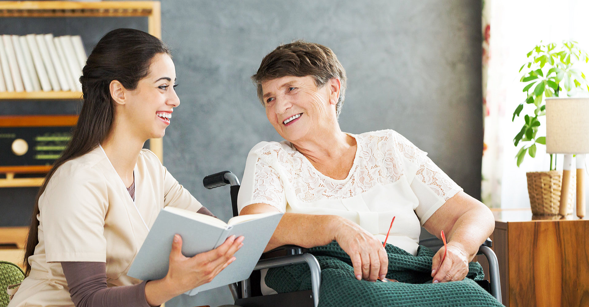caregiver holding a book and senior woman with wheelchair are smiling